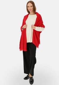 Czerwony szal zarzucony na kremowy sweter, noszony z czarnymi szerokimi spodniami i błyszczącymi czarnymi butami. Widoczne szczegóły frędzli na krawędziach szala.