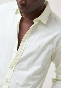 Camisa de botón amarilla claro hecha de una tela suave, con un cuello clásico y un logotipo circular en el pecho. Costuras limpias y botones visibles.