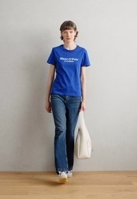 Modré bavlněné tričko s bílým textem, spojené s tmavými džíny. Modelka drží bílou tašku, stojí na dřevěné podlaze s šedou zdí v pozadí.