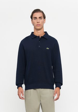 Man wearing navy long-sleeve polo with green crocodile logo and beige pants, standing against plain white background.
