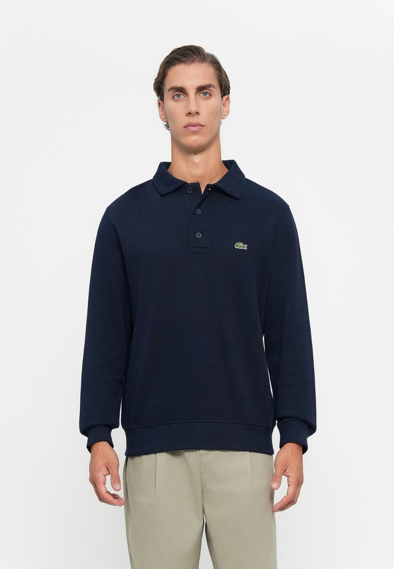 Mann, der ein navyblaues Langarm-Polo mit grünem Krokodil-Logo und beigen Hosen trägt, steht vor einem schlichten weißen Hintergrund.