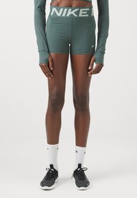 Zöld sportnadrág, "NIKE" logóval a derékrészen, hosszú ujjú felsővel és fekete futócipővel, fehér zoknikkal kiegészítve.