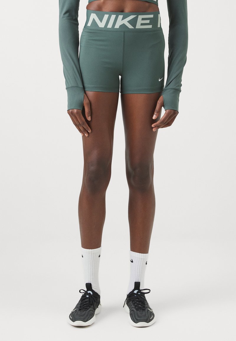 Shorts de sport verts avec le logo "NIKE" sur la taille, associés à un haut à manches longues et des chaussures de course noires, avec des chaussettes blanches.