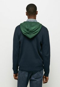 Marinblå sweatshirt med grön huva, som har en mjuk textur, långa ärmar och ribbade muddar. Avslappnad design med accentstickningar.