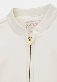 Jachetă bomber din piele albă cu guler rotund, prevăzută cu un fermoar auriu cu pandantiv cu logo și o etichetă de brand în interiorul gulerului.