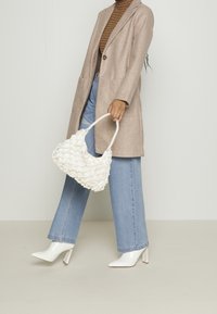 Žena v béžovém kabátu, pruhovaném roláku, světle modrých džínech a bílých kožených kotníkových botách, která drží bílou síťovanou kabelku před jednolitým pozadím.