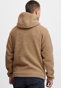 Tansfarvet fleece-hoodie med en tekstureret overflade, der har hætte og lange ærmer. Sømdetaljer løber langs skuldre og sider.