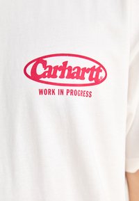 Camiseta de algodón blanca con un logotipo ovalado rojo que incluye el texto "Carhartt" y "WORK IN PROGRESS" en una fuente negrita y redondeada. Mangas cortas.