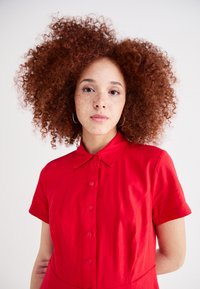 Κόκκινο φόρεμα με κουμπιά και κοντά μανίκια, γιακά και ορατά κουμπιά. Σγουρά μαλλιά και δέρμα με φακίδες. Απλός, καθαρός σχεδιασμός σε ουδέτερο φόντο.