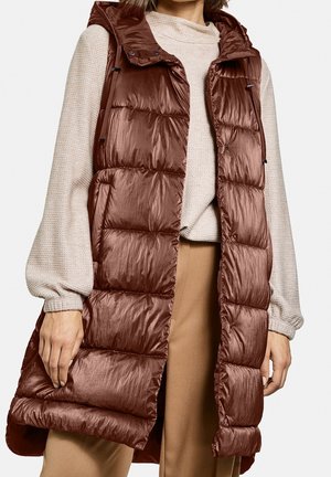 Gilet sans manches marron et rembourré avec une texture brillante, une capuche et des détails à cordons, porté sur un pull léger et un pantalon beige.