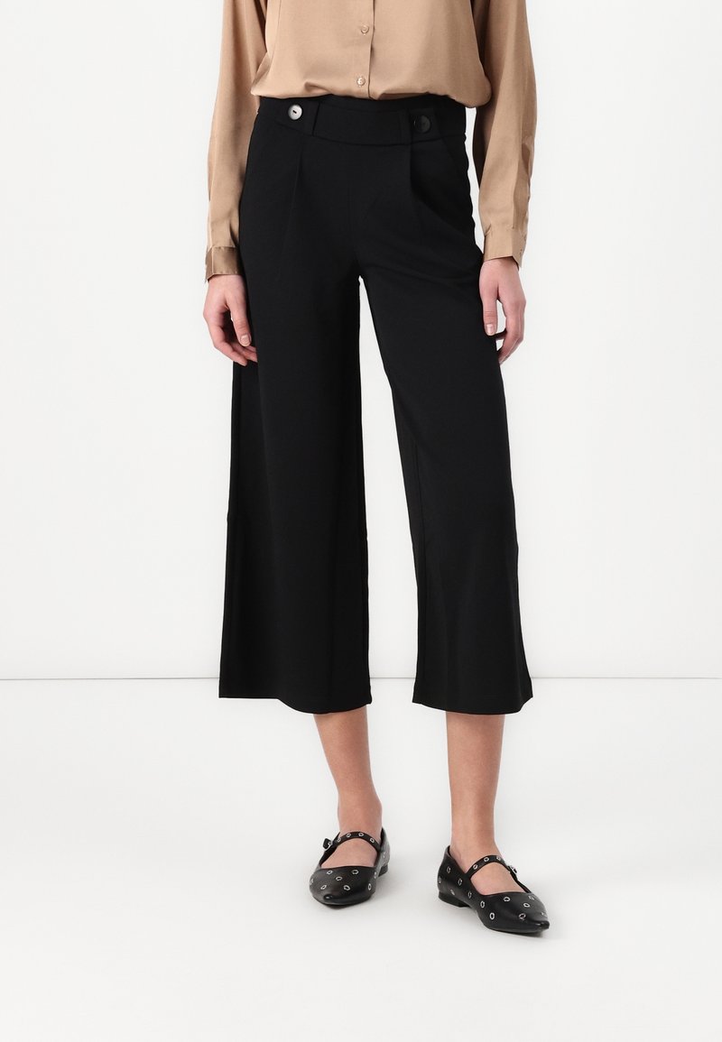 Pantalon noir raccourci à taille haute, avec deux plis avant et détails de poches. Tissu à finition lisse, assorti à des chaussures plates noires.