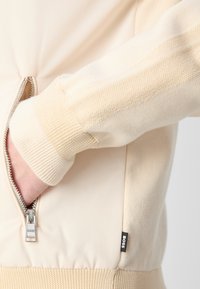 Beige zip-up jacka med texturerade stickade ärmar, ribbade manschetter och en synlig logotag. Slät tyg med en figurnära design.