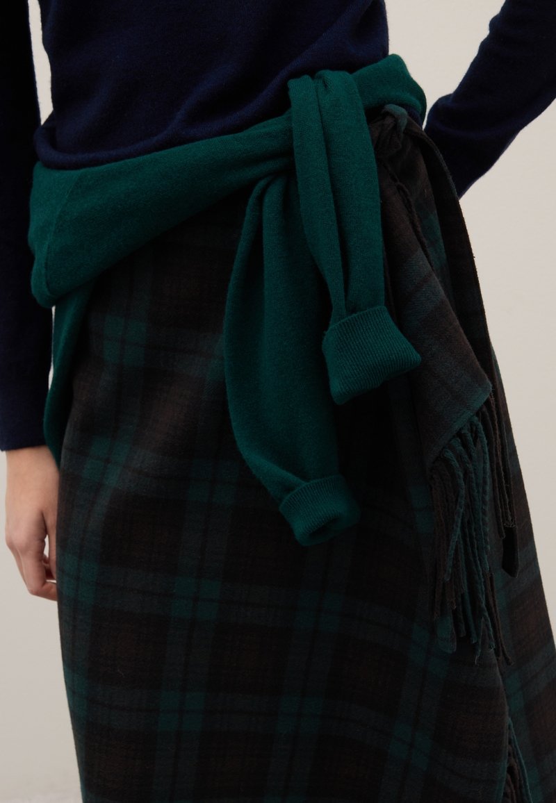 Pull vert en tricot noué autour d'une jupe à carreaux noirs et verts avec des bords à franges. Le tissu semble doux, avec un aspect superposé.