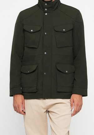 Dunkelgrüne Jacke mit vier Klappentaschen und silbernen Druckknöpfen, getragen über beige Hose, zeigt Vorderseite des Oberkörpers und Arme.