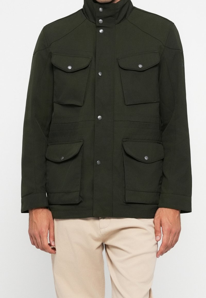 Veste vert foncé avec quatre poches à rabat et boutons-pression argentés portée sur un pantalon beige, montrant le buste et les bras de face.