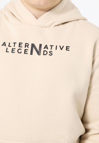 Felpa beige realizzata in tessuto morbido, con una grande stampa nera della parola "ALTERNATIVE" sul petto e un cappuccio con cordino.