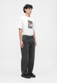 Joven con cabello rizado que lleva una camiseta gráfica blanca con una estampación de un perro, jeans grises holgados y zapatillas negras, de pie de lado.