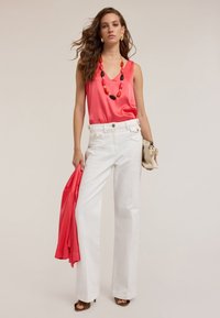 Top sin mangas de satén coral combinado con pantalones blancos de cintura alta. La modelo sostiene un chal rosa, lleva un collar de cuentas multicolores y tacones marrones.