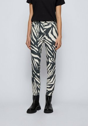 Personne portant un t-shirt noir à manches courtes, un pantalon imprimé zèbre noir et blanc, et des bottines noires épaisses, debout devant un fond gris uni.