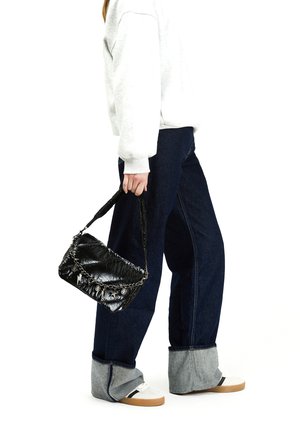 Borsa nera e gonfia con tracolla a catena, decorata con dettagli metallici, tenuta da una persona che indossa un felpa grigia e jeans blu oversize con orli risvoltati.