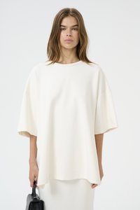Üks suurus lühikeste varrukatega topp pehmes valges kangas, ümmarguse kaelusega ja diskreetse reljeefse logoga, kantud koos musta käekotiga.
