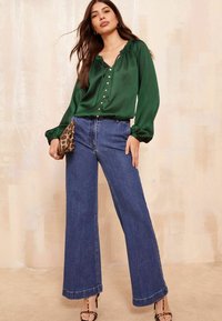 Grüne Satinbluse mit V-Ausschnitt, Knopfakzenten und gerafften Ärmeln, kombiniert mit hoch taillierten, weit geschnittenen blauen Jeans.