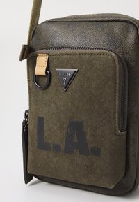 Grønn lerrets crossover-veske med en teksturert brun topp, med svart "L.A." trykk, en Guess-logo og en glidelås hovedkompartment.