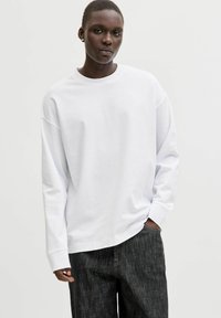 Hvid langærmet t-shirt lavet af blødt stof med en afslappet pasform, rund halsudskæring og sænkede skuldre, kombineret med mørke teksturerede jeans.