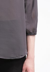 Grå gennemsigtig bluse med trekvartslange ærmer, elastiske manchetter og et blødt, letvægtsstof. Designet har en glat overflade.