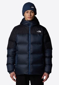 Granatowa kurtka puchowa z czarnym kapturem, zapinana na zamek, z kieszeniami na suwak i ściągaczami przy mankietach. Posiada logo na piersi i wykorzystuje elegancki, pikowany design.