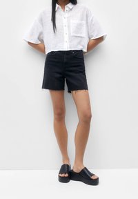 Chemise blanche à manches courtes avec boutons et poche, associée à un short en denim noir taille haute et des sandales noires à plateforme.