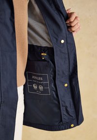 Person hält eine offene marineblaue Jacke, die eine gebrandete Innentasche mit der Aufschrift „Joules Right As Rain“ mit wasserabweisenden und winddichten Symbolen zeigt.