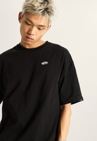 Camiseta de algodón negra con un pequeño logo blanco de "Vans" en el pecho, mangas cortas y escote redondo. Diseño simple y discreto.