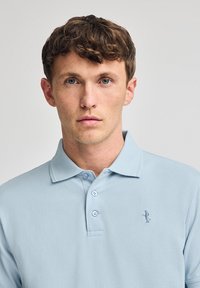 Polo de color azul claro, hecho de algodón, con cuello, tres botones y un pequeño logotipo bordado en el lado izquierdo del pecho. Textura suave.