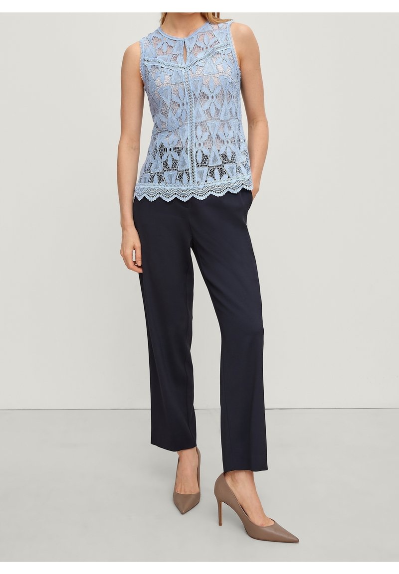 Femme portant un top en dentelle bleu clair sans manches, un pantalon noir droit et des chaussures à talons taupe, debout avec une main dans la poche.