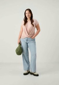 Νεαρή γυναίκα με μακριά μαλλιά, φορώντας �ροζ μπλούζα με πλέξη, ανοιχτό μπλε παντελόνι με φαρδιές γραμμές, λευκές μπότες και κρατώντας μια πράσινη τσάντα με καπιτονέ σχέδιο.