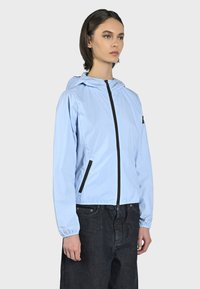 Giacca a vento blu chiaro con cappuccio, cerniera frontale e dettagli neri; presenta polsini elasticizzati e tasche laterali. Indossata sopra jeans scuri.