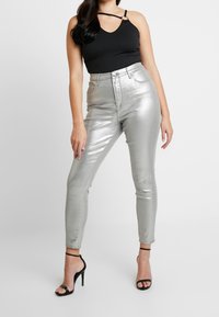 Pantalones altos de metal plateado con un ajuste ceñido, textura suave y acabado brillante, combinados con una camiseta sin mangas negra y tacones negros con tiras.