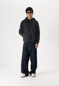 Un sweat à capuche gris foncé avec une poche avant et un cordon, associé à un jean bleu foncé traditionnel et des baskets blanches. Des lunettes de soleil noires.
