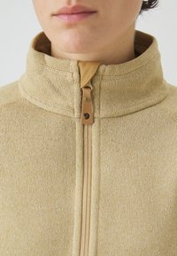 Fleece beige con zip e colletto alto, caratterizzato da una trama liscia e un accento in pelle marrone vicino alla zip.
