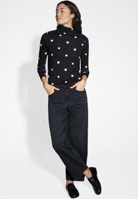 Zwarte lange mouw top met witte stippen, gecombineerd met donkere spijkerbroek. De top heeft een hoge kraag; de schoenen zijn zwart en plat.