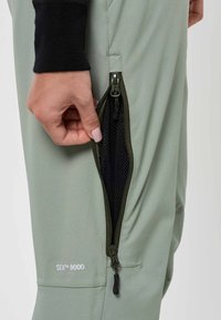 Hand die de verticale zijrits opent van lichtgroene broek met zwarte mesh voering en STX 8000-logo op het been.