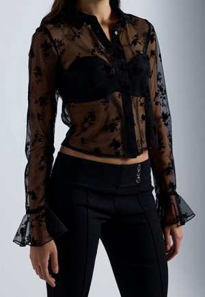 Femme portant un chemisier noir transparent à manches longues avec des motifs floraux sur un soutien-gorge noir, associé à un pantalon noir taille haute avec trois boutons.