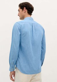Lichtblauw denim shirt met lange mouwen, knoopmanchetten en een rugplooi. Soepele textuur met een iets getailleerde pasvorm.