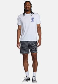 Wit T-shirt met blauwe grafische print op de borst, gecombineerd met zwarte patroon shorts met schedelontwerp. Zwarte sneakers en witte sokken.