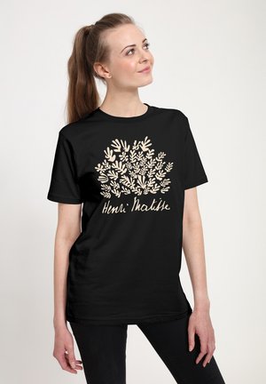 Ung kvinde med en sort Henri Matisse t-shirt med beige abstrakte bladformer, der står og kigger op til siden.