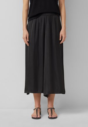 Kobieta ubrana w czarne, szerokie, krótkie spodnie z marszczonym paskiem i czarne sandały odkryte na palcach, stoi na szarym tle.