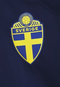 Emblema em forma de escudo azul com detalhes amarelos. Apresenta um ícone de bola de futebol e o texto "SVERIGE", sobre um fundo de tecido azul escuro.