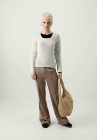 Sieviete ar īsiem blondiem matiem, tērpusies baltā džemperī, brūnos platos biksēs, melnās kurpēs un tur lielu beigtu ķīļveida somu.