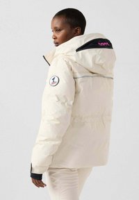 Giacca a puff panna con cappuccio, caratterizzata da accenti neri, un logo rotondo e dettagli colorati sulla schiena. Materiale strutturato e isolante.